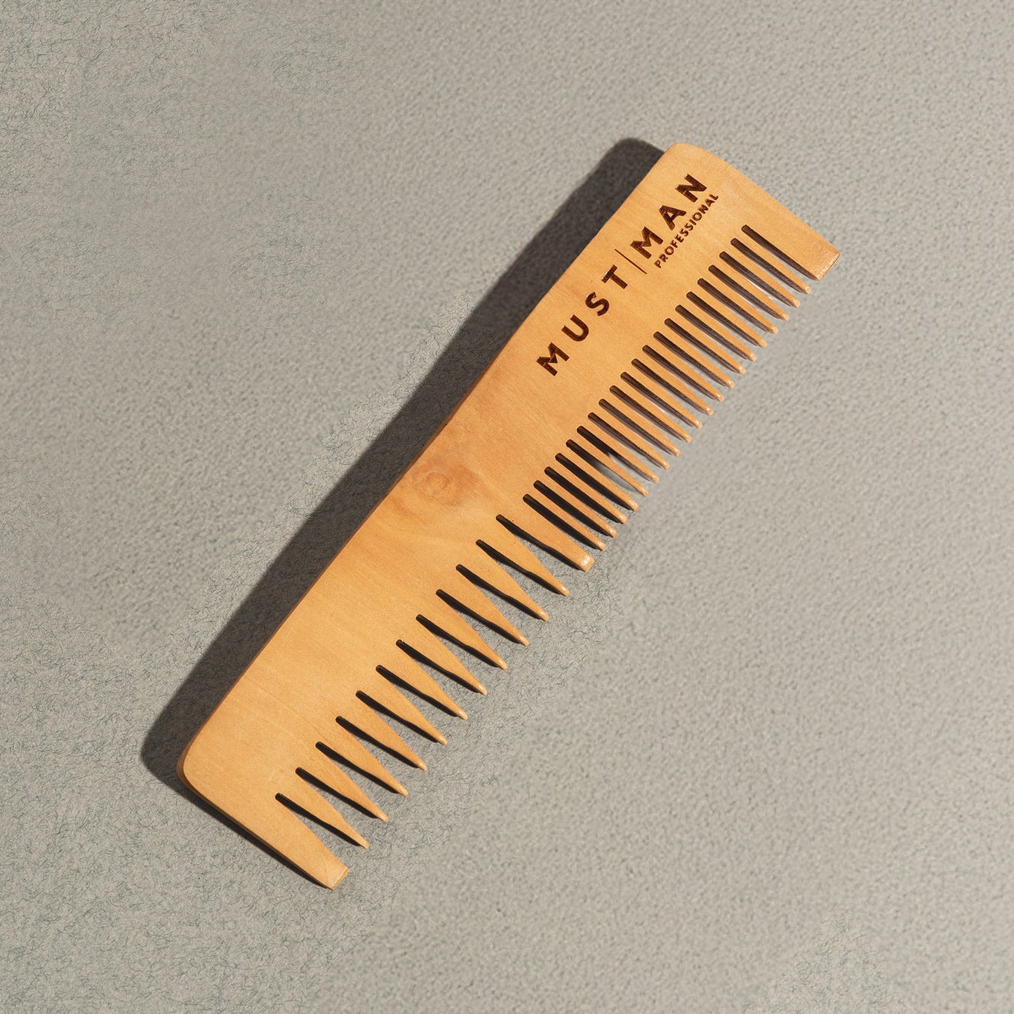Must Man Professional Styling Hero Comb — peach wood comb angled on a grey surface, showing the dual tooth spacing
