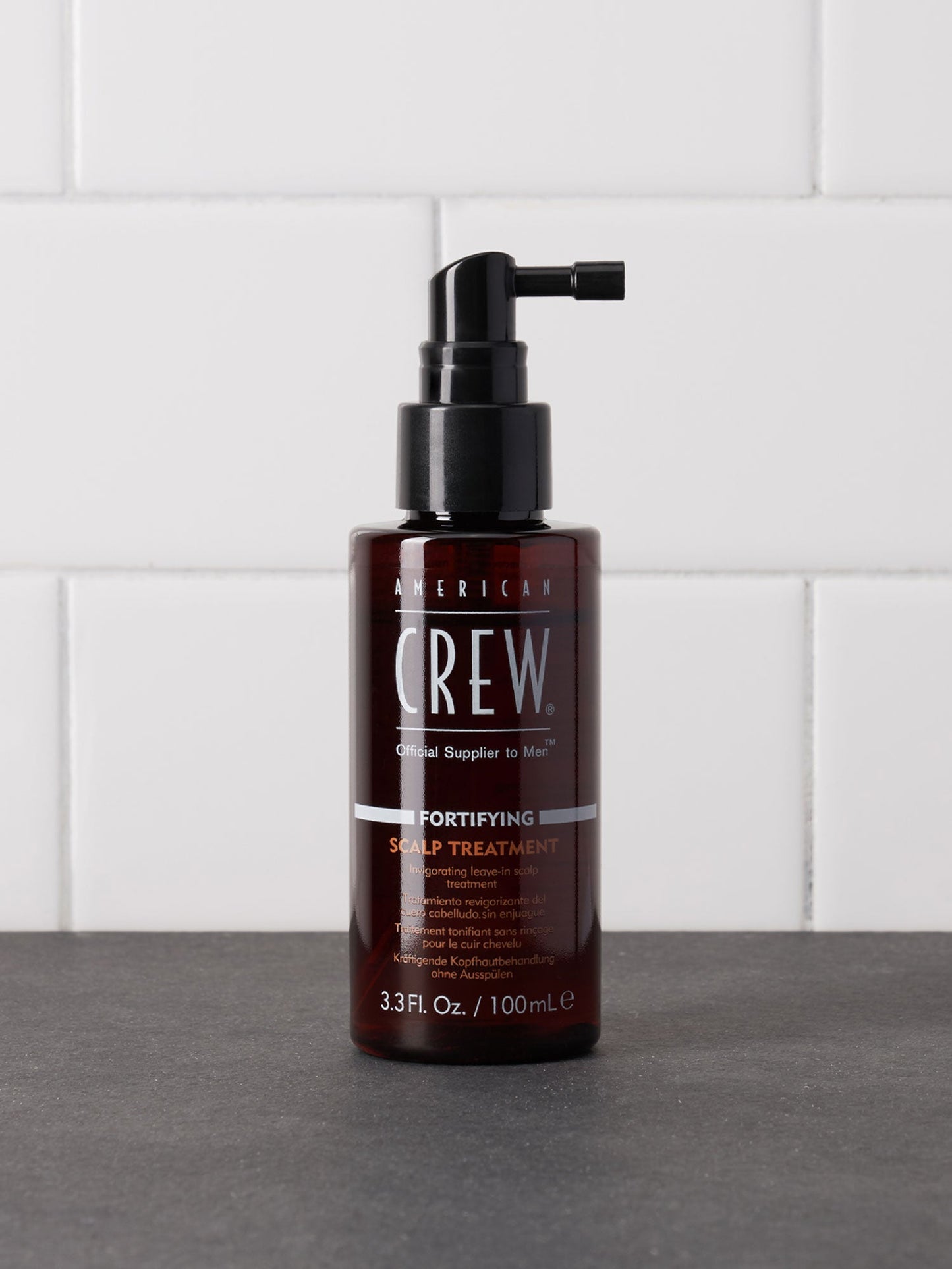 American Crew Fortifying Scalp Treatment — 100 ml spray bottle standing on a dark counter against white tiles, label reading 3.3 Fl. Oz. / 100 mL