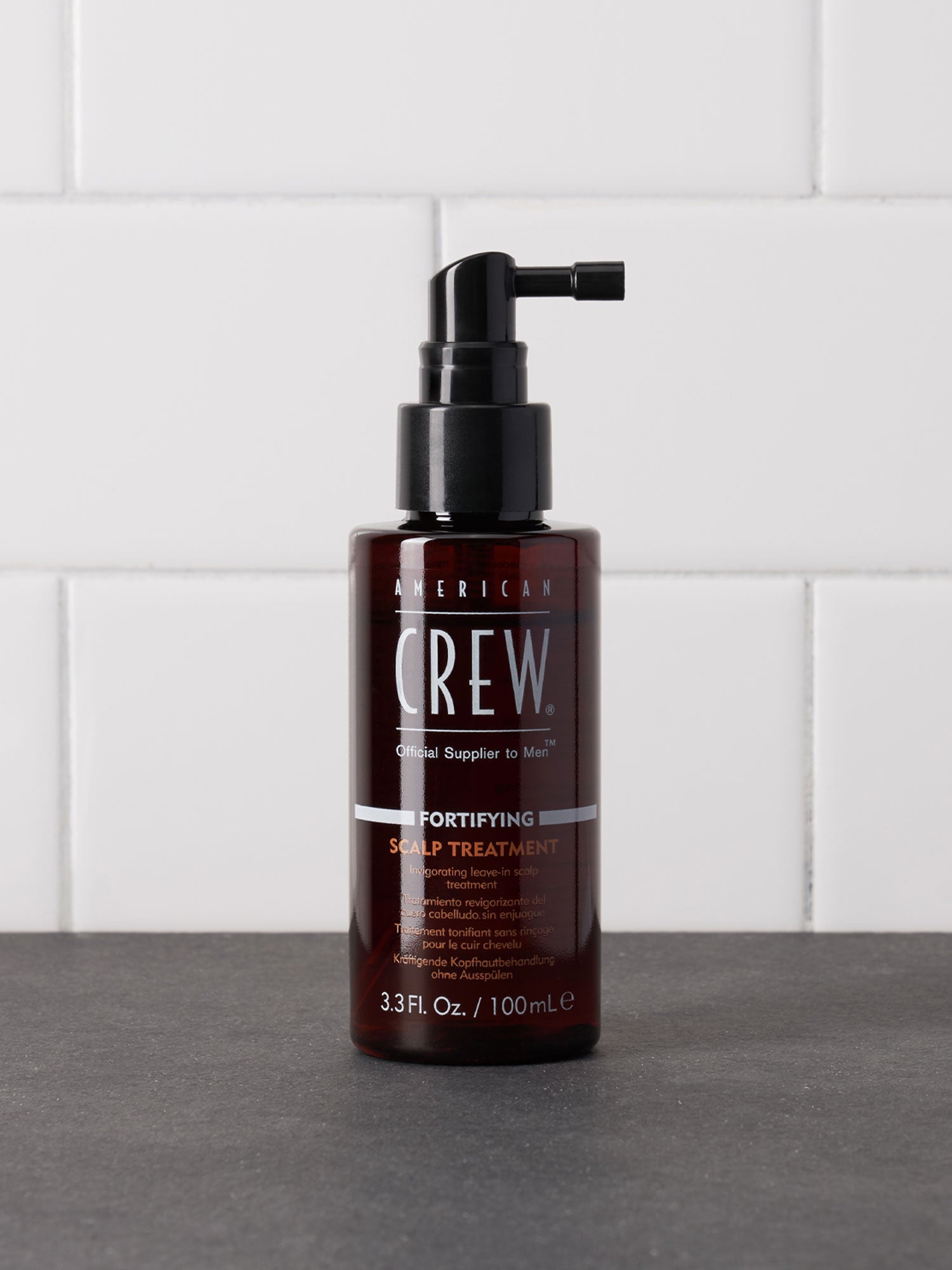 American Crew Fortifying Scalp Treatment — 100 ml spray bottle standing on a dark counter against white tiles, label reading 3.3 Fl. Oz. / 100 mL