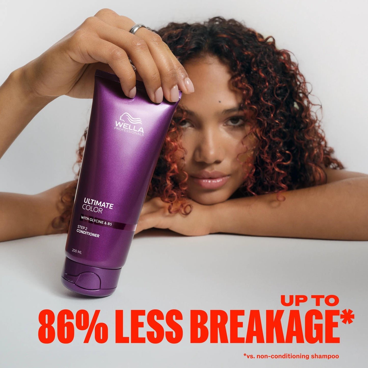 Wella Professionals Ultimate Color — Image of model with curly brunette hair and red streaks, leaning on their chin with one hand and holding the Ultimate Color conditioner with their other. Text overlay reads "Up to 86% less breakage* *vs non-conditioning shampoo".